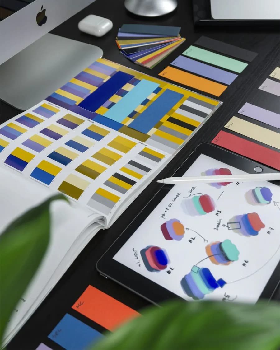 Desk with Pantone-style color swatch books, an iPad showing brand color mockups, and an iMac